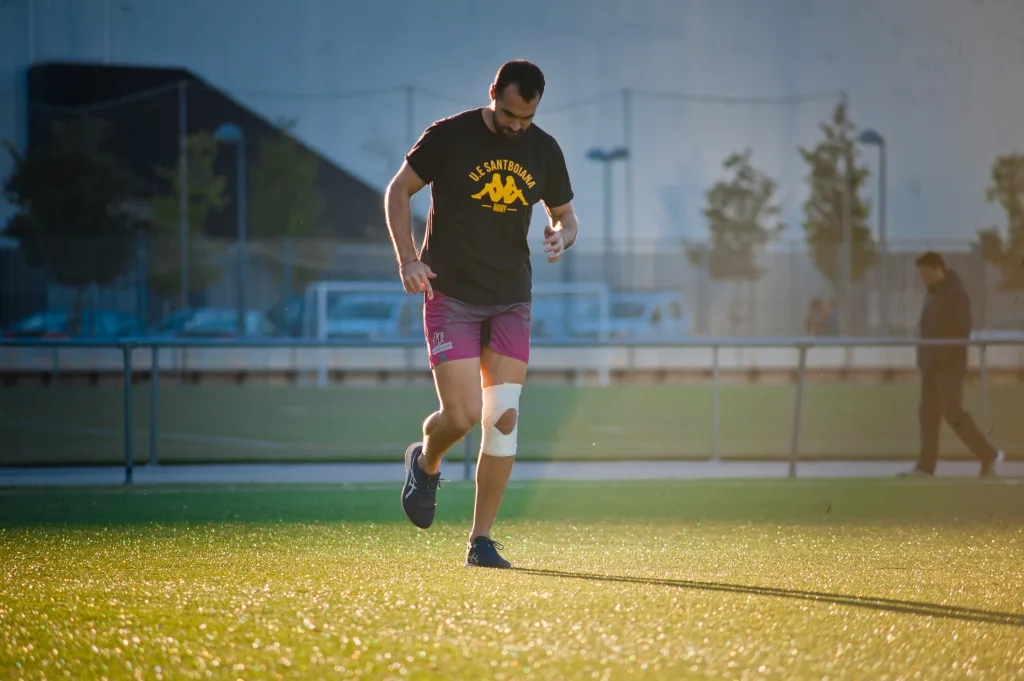 Atleta con vendaje en la rodilla corre en un campo de césped sintético al atardecer, entrenando su movilidad tras una lesión.